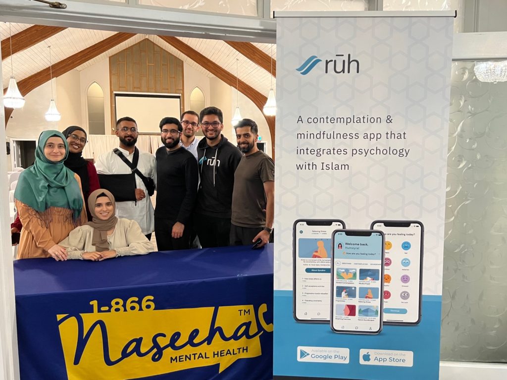 A group of Ruh team members gathered behind a table, some sitting, some standing. To their left is a large pop up retractable banner for Ruh. On it, there is the Ruh logo, text that reads "A contemplation & mindfulness app that integrates psychology with Islam," and images of a phones displaying different pages within the app.