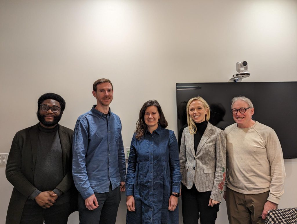 The 5 judges stand together in front of a white wall, smiling at the camera.