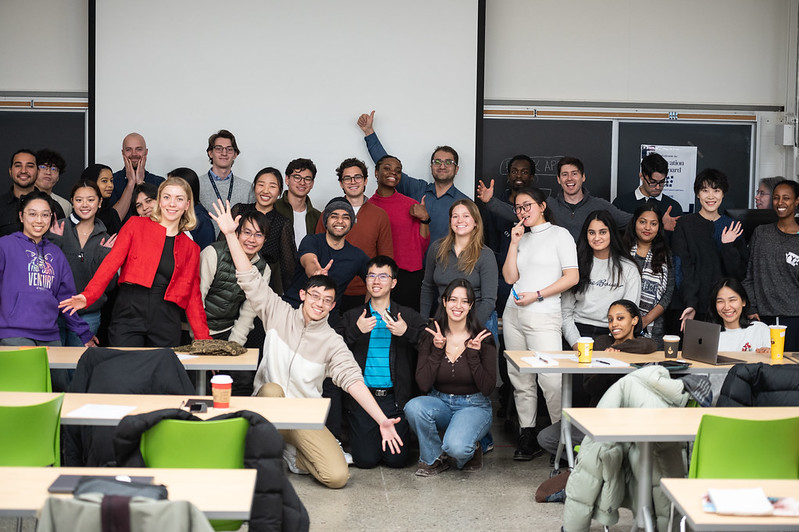 Group of participants in Co-Founder's day gather for a group shot at the front a classroom, smiling, showing peace signs and other celebratory gestures